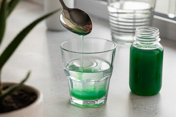 Aloe gel flows from a spoon into a glass of water, against the background of an aloe flower and a bottle of healing gel. The concept of using natural plants in the treatment of stomach diseases.