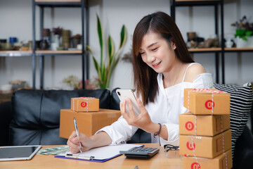 Woman are receiving orders via smartphone. Business woman owner holding parcel box for delivery shipping and using smartphone at home office. Business owner talking on mobile phone and taking order.