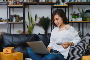 Asian woman holding a credit card while shopping online with a laptop at home. Concept of e-commerce, lifestyle, and digital payment in a cozy modern living space.