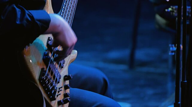 A musician's hand skillfully plays a bass guitar, showcasing intricate finger movements. The scene is illuminated with a blue hue, creating an engaging atmosphere