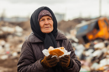 Old homeless woman holds bread in her hands against the background of a garbage dump