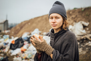 A young homeless woman holds bread in her hands against the background of a garbage dump