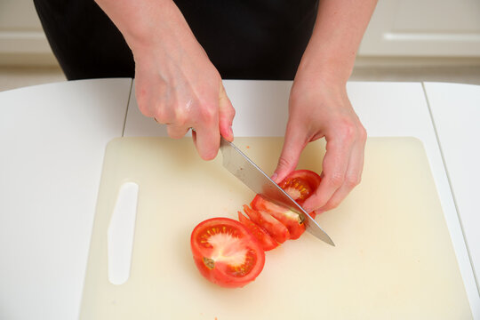 Close-up of hands cutting a ripe tomato on a plastic cutting board with a kitchen knife, preparing for cooking or salad.
