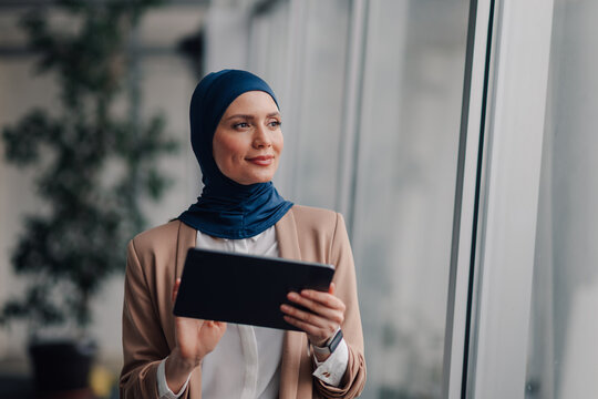 Businesswoman wearing hijab holding tablet and looking out window - Powered by Adobe