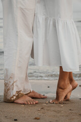Romantic barefoot moment on the beach. Couple in white clothes stands close in soft evening light. Warm, natural and intimate coastal scene.