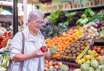 Elderly woman holding a red tomato and a green artichoke while buying fresh vegetables at the market, choosing from a wide selection. Healthy eating, the concept of shopping and consumerism