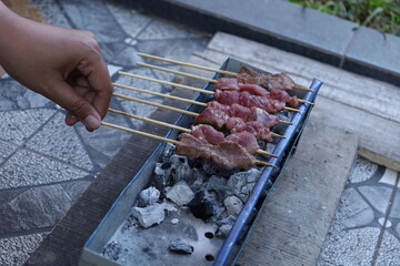 A woman's hand holds a skewer. Goat satay is being grilled over hot coals. Fresh meat is being grilled using a wooden skewer over hot coals. Side view photo.