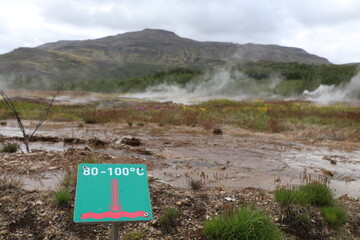 volcanic landscape in Geysir, Iceland with steam coming off hot water in different hydrothermal springs, a sign warns for the high water termperature