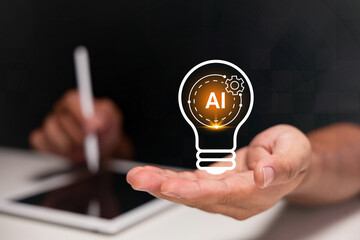 A hand holds a glowing AI lightbulb icon, with a person using a tablet and stylus in the background. © Minttita