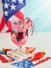 4th of July Holiday background. Punch with ice and strawberry pieces in glass and  beautiful cocktail straw with fireworks against background of old turquoise board and sun rays.