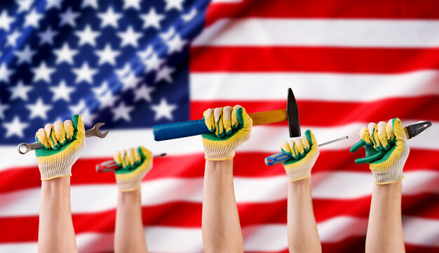 Hands of mans workers holds different tools hammer, wrench, screwdriver, pliers in gloves against background of American flag. Labor day concept. Holiday background.