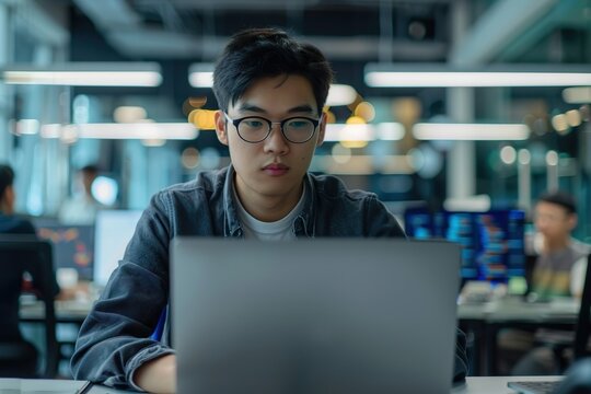 A young Asian male software engineer is seen intensely focused on his laptop screen, coding in a modern tech office. Wearing glasses and a plaid shirt, he types with concentration while colorful lines