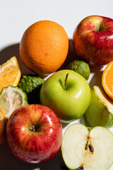 Assortment of fresh fruits including green apples, red apples, oranges, and kaffir limes arranged on a white background with strong shadows and bright lighting emphasizing texture and freshness.