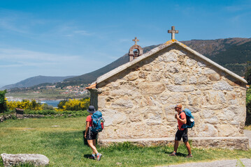 Couple of pilgrims with backpacks walking next to old church on Camino Portuguese Way, inspiring...