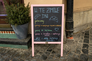 Obraz premium A charming cafe chalkboard with handwritten Polish text stands on a cobbled street next to a festive evergreen plant. The board promotes winter specials, creating a cozy seasonal atmosphere