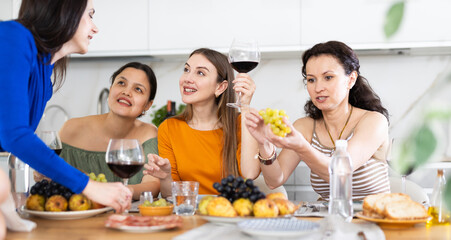 Relaxed adult female friends having fun at intimate house party, sharing stories and laughing gathered around table with wine and appetizers in cozy kitchen