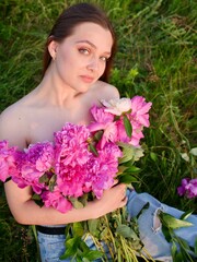 Happy smiling young woman in summer outdoors in a field with pink peony flowers enjoying life in a moment at sunset