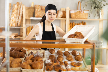 Shopping area, showcase, counter in bakery. Girl employee with long hair fills window and puts croissants on counter shelf, puts buns on shelf, shows and demonstrates wide selection of pastries