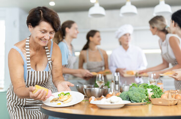 Mature woman decorating cooked chicken breast with mustard sauce posing surrounded by other members of cooking course