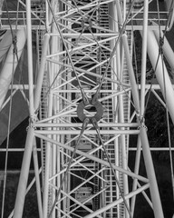 Black and white photograph capturing the structural details of a Ferris wheel in Riga, Latvia. The image highlights the intricate geometry and contrast of the metal framework against the sky. 