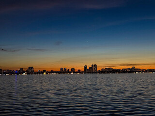 Naklejka premium Silhouetted skyline along the waterfront at sunset with city lights reflecting on the water in a tranquil setting