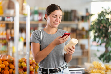 Fototapeta premium European woman scans the barcode of a package with mayonnaise for contactless payment at the checkout. Customer pays online for groceries in a supermarket using a qr code
