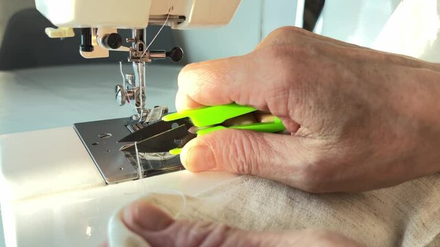 Close-up of an experienced seamstress cutting fabric with green scissors near a sewing machine. The pensioner's hands are busy with work 