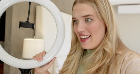 Female vlogger greeting viewers, removing phone mount, showing ring light setup at home, copy space