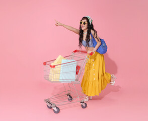 Happy smiling young asian woman dressed in summer clothes pushing trolley cart and holding shopping bag summer sale isolated on pink background.