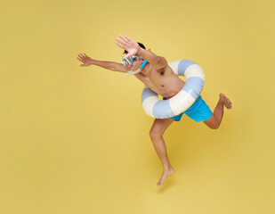 Full length cheerful young shirtless man in swim snorkel and inflatable ring jumping to dive having fun isolated on yellow copy space background.
