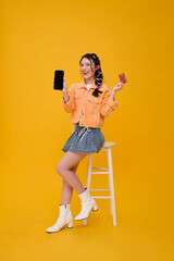Happy cute young Asian teen woman using mobile phone and showing credit card payment for shopping online. While her sitting on chair isolated on yellow background.