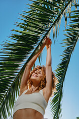 smiling woman with curly hair reaches up to green palm leaves under clear blue sky, wearing casual white top, sunlight highlighting joyful summer mood.