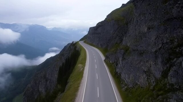 Top-down view of a narrow mountain road snaking along a ridge with dramatic cliffs on both sides and low-hanging clouds drifting by. The peaks in the distance fade into mist, and the road remains pris