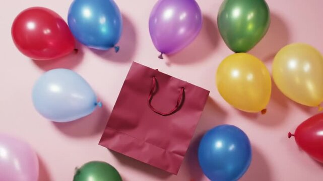 Colorful balloons scattered around a maroon gift bag on a pink surface in a celebratory arrangement