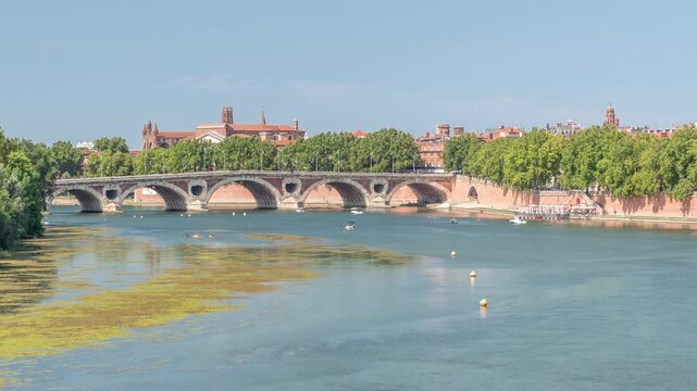 Garonne River and Pont Neuf timelapse in downtown Toulouse, France. Renaissance arch bridge reflects in the water under a blue sky with Basilica of Our Lady of the Daurade. Waterfront with green trees