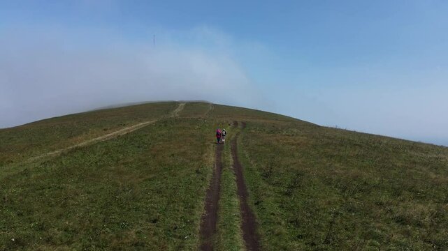A drone rises from the ground, filming a man and a woman &mdash; hikers with backpacks &mdash; walking away on a mountain dirt road against a blue sky.