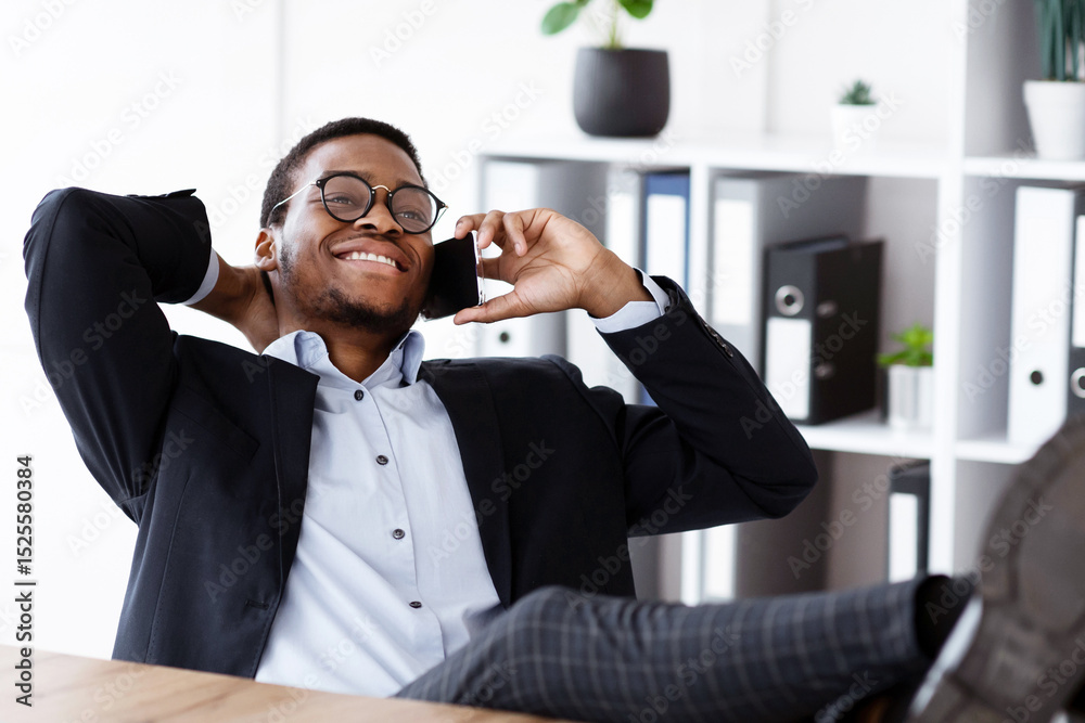 Poster relaxed black manager sitting with legs on workdesk, having conversation on mobile phone and smiling
