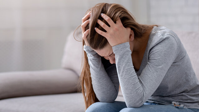 Unhappy young woman with head in her hands sitting on couch, crying and feeling depressed. Desperate millennial lady suffering from mental problems, broken heart or mood disorder