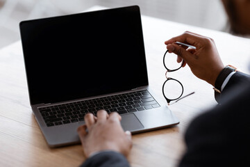 Unrecognizable black businessman using laptop with blank screen, holding glasses, shot over...