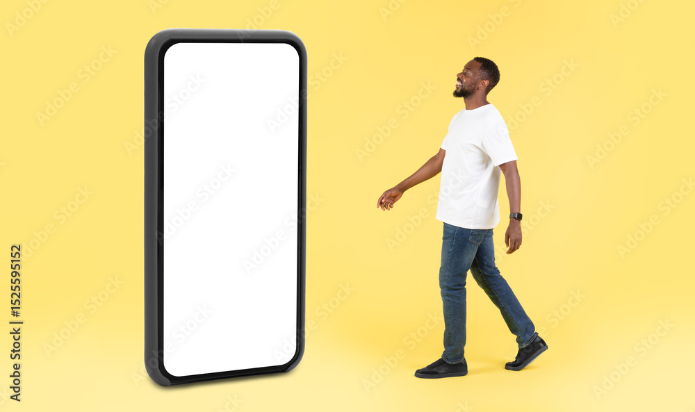 Poster black guy walking near huge phone with empty screen in studio over yellow background. great mobile a