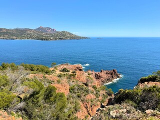 Scenic coastal landscape in the Esterel region of the French Riviera with red cliffs, pine trees, and a clear blue sea stretching toward the horizon under a bright sky.
