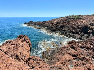 Dramatic Mediterranean coastline with red and dark volcanic rocks and turquoise water under a clear blue sky, captured on a sunny summer day in the Esterel region, southern France.
