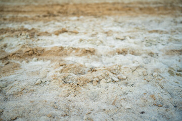 A close-up shot of textured sand, mixed with some small rocks and lighter material, fills the frame. The focus is on the details of the sands surface.