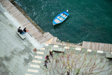 Obraz premium Aerial view of a blue rowboat moored on the Mediterranean coast, with people walking and sitting on a stone promenade. Captures a peaceful seaside moment in an Italian village