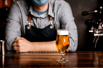 Quarantine, nightlife and pub. Bartender in protective mask leans on bar counter on which stands glass of light beer with foam, cropped