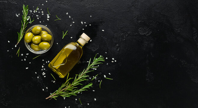 Olive oil, fresh olives and rosemary sprigs on black background, top view, copy space. Organic olive oil concept