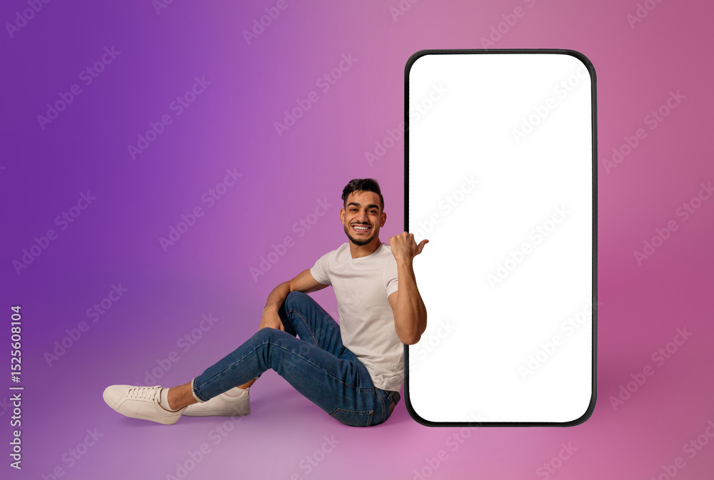 Poster full length of cheerful young arab man sitting near big cellphone with mockup for mobile app on scre