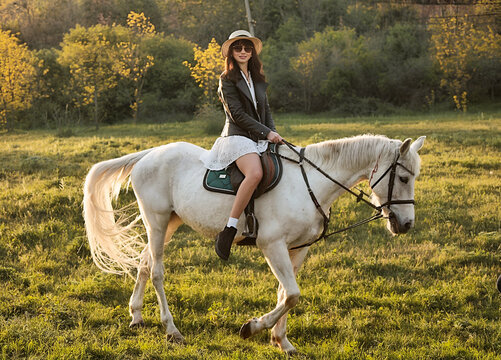 Beautiful young girl with dark hair on a white horse in a field at sunset in spring