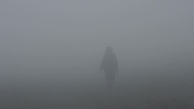 Female hiker walks away on a mountain road in thick fog and clouds. Strong wind, low visibility, harsh weather conditions.
