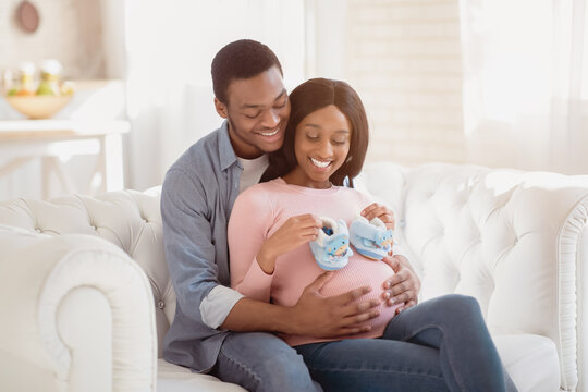 Portrait of positive pregnant couple with tiny baby booties hugging on couch indoors. Romantic expectant woman and her caring husband cuddling together on sofa, expressing love for each other
